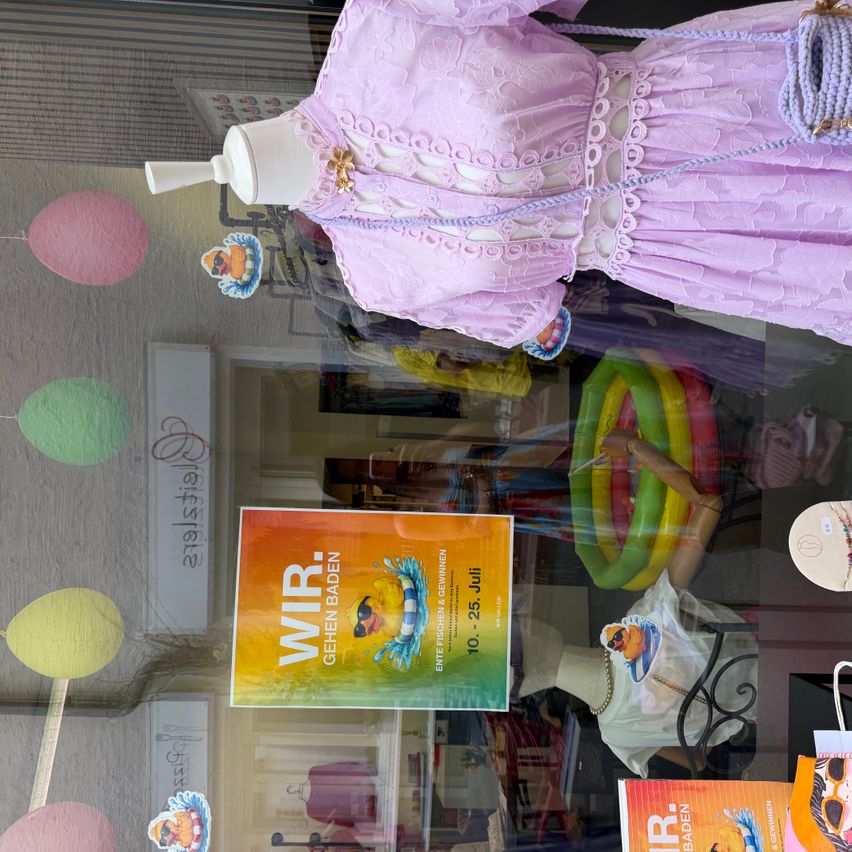 Ein Mannequin in einem rosa Spitzenkleid wird in einem Schaufenster ausgestellt. Ein Plakat mit der Aufschrift 'Wir Gehen Baden' ist im Vordergrund sichtbar.