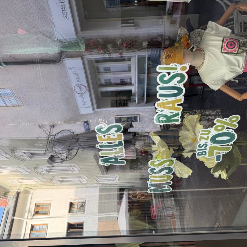 Ein Schaufenster zeigt ein Mannequin, das einen Hut und ein T-Shirt mit einem bunten Design trägt. Das Fenster hat auch Aufkleber mit den Worten 'Ales' und 'Miss Praus' und einer gelben Blume. Die Reflexion des Gebäudes und des Mannequins ist auf dem Glas zu sehen.