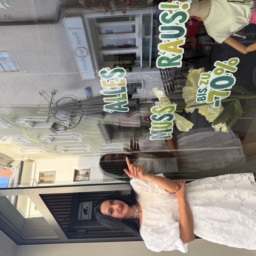 Eine Frau in einem weißen Kleid posiert vor einem Schaufenster mit Schildern, die einen Verkauf bewerben, und einer Statue im Hintergrund.