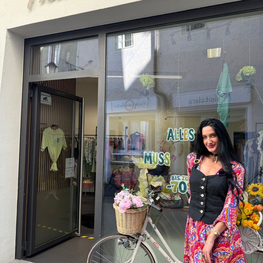 Eine Frau steht vor einem Laden mit einem Fahrrad und einem Korb mit Blumen. Das Schild des Ladens lautet Alles Muss.