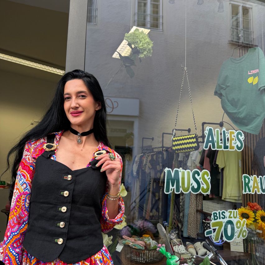 Eine Frau mit langen schwarzen Haaren, die eine bunte Weste und eine schwarze Kette trägt, steht vor dem Schaufenster eines Bekleidungsgeschäfts mit einem Verkaufsschild, das 'Alles muss raus' und 'Bis zu -70%' anzeigt.