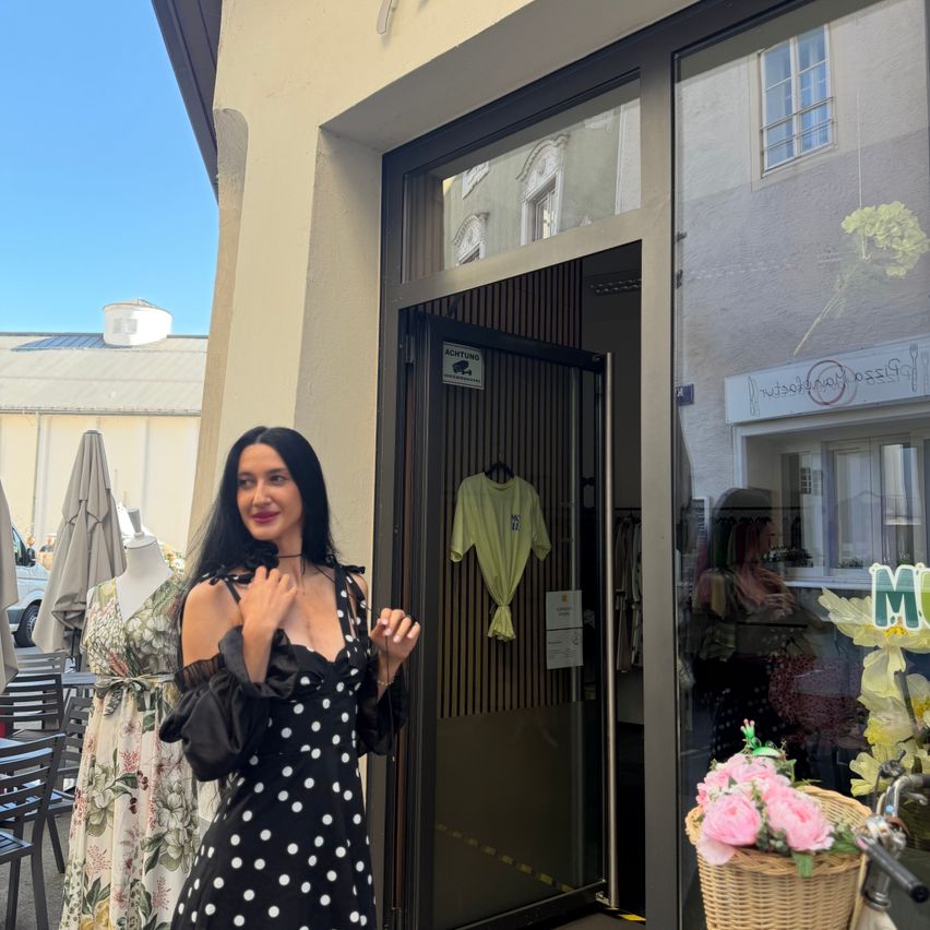 Eine Frau in einem gepunkteten Kleid steht vor einem Boutique mit einem gelben Blumenarrangement in einem Korb.