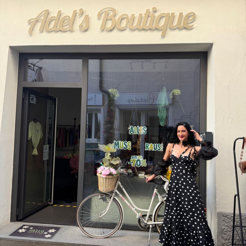 Eine Frau in einem gepunkteten Kleid steht vor Adel's Boutique und hält ein Fahrrad mit einem Korb voller Blumen. Die Glastür des Ladens spiegelt ein Verkaufsschild und Innendisplays wider.