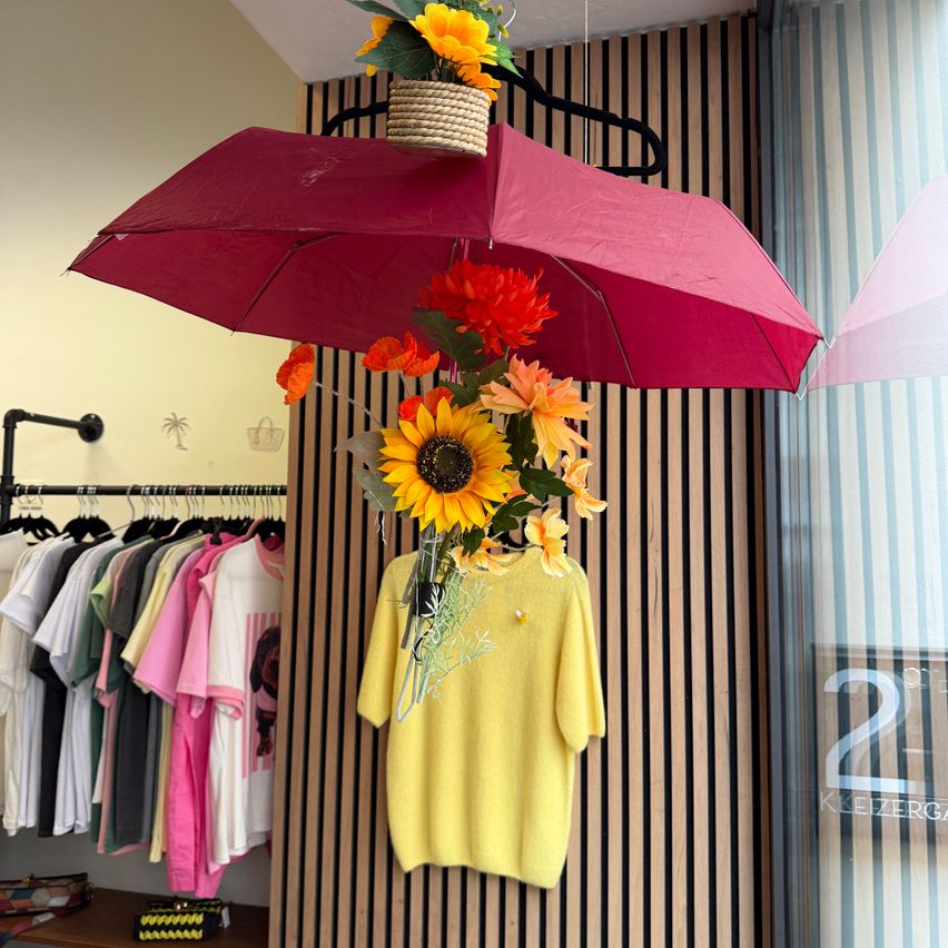 Ein gelbes Sweatshirt mit einer Blumenarrangement aus Sonnenblumen und Gänseblümchen hängt von einem roten Regenschirm in einem Bekleidungsgeschäft.