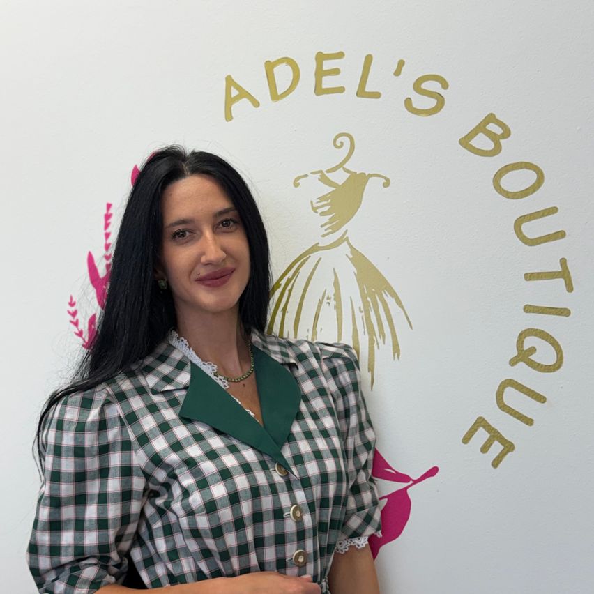 Eine Frau mit langen schwarzen Haaren, die eine grün-weiß karierte Bluse trägt, steht vor einer weißen Wand mit einem goldenen Logo, das 'ADEL'S BOUTIQUE' liest.