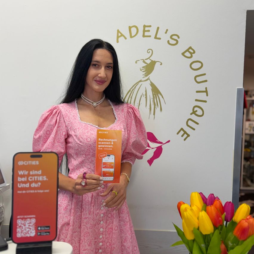 Eine Frau in einem rosa Kleid steht vor einer Wand mit dem Logo von Adel's Boutique, hält ein Flugblatt und ein Smartphone in der Hand. Sie lächelt und scheint für ein Foto zu posieren.