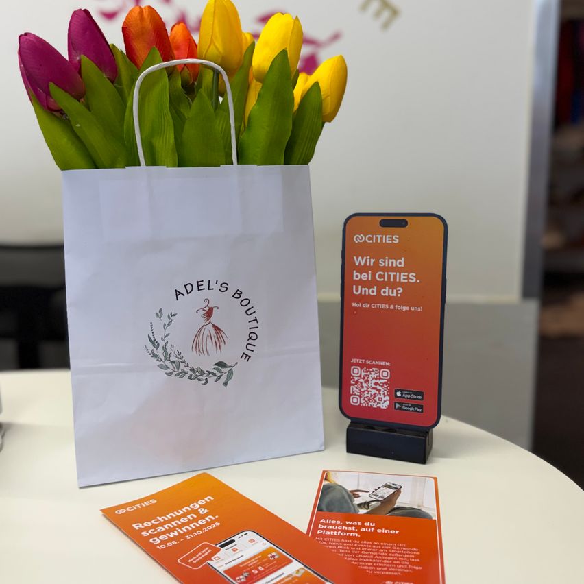 Ein weißer Papierbeutel mit Adel's Boutique Logo, der einen Strauß mehrfarbiger Tulpen enthält, neben einem roten Telefonständer mit QR-Code und einem Flyer.