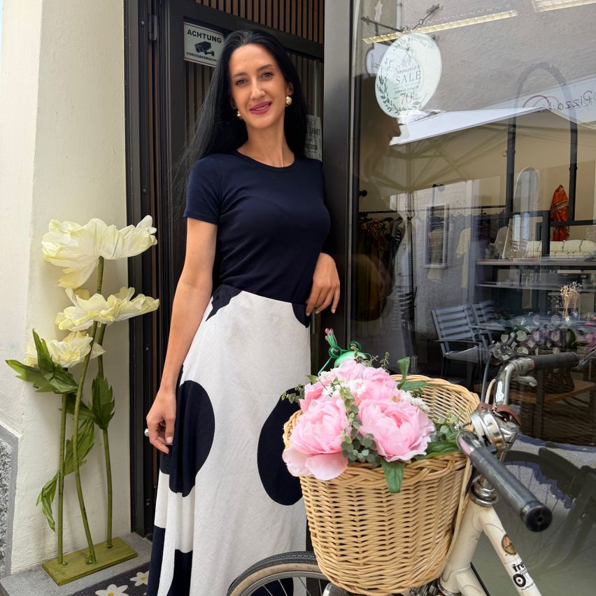 Eine Frau steht vor einem Geschäft mit einem Fahrrad und Blumen. Sie trägt einen schwarz-weiß gepunkteten Rock und ein marineblaues Oberteil.