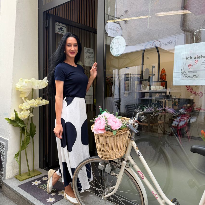 Eine Frau in einem gepunkteten Kleid steht vor einem Geschäft mit einem Fahrrad und einem Korb voller Blumen.
