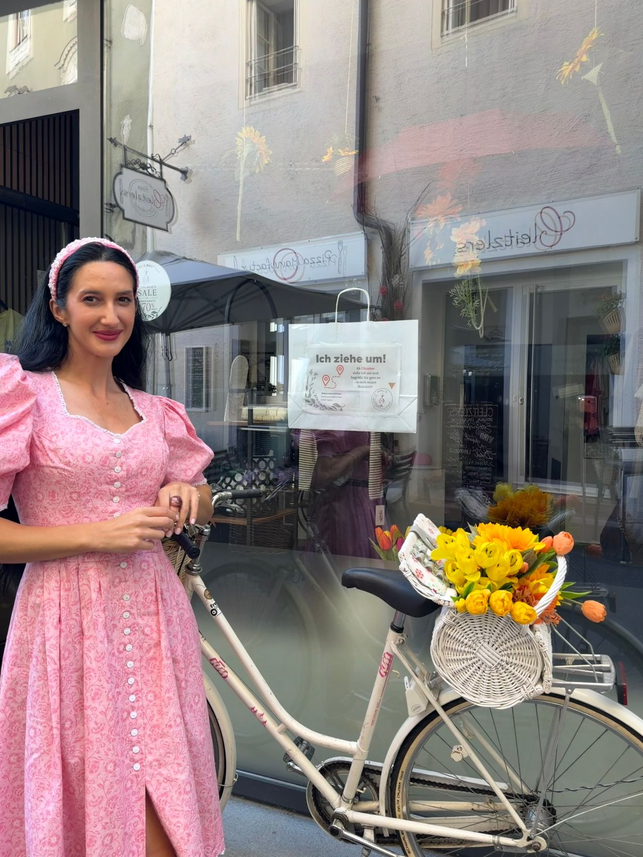 Eine Frau in einem rosa Kleid steht neben einem Fahrrad mit einem Korb gelber Blumen. Hinter ihr befindet sich ein Geschäft mit einem Schild, das 'Ich ziehe um!' besagt, und einer Reflexion einer Person.