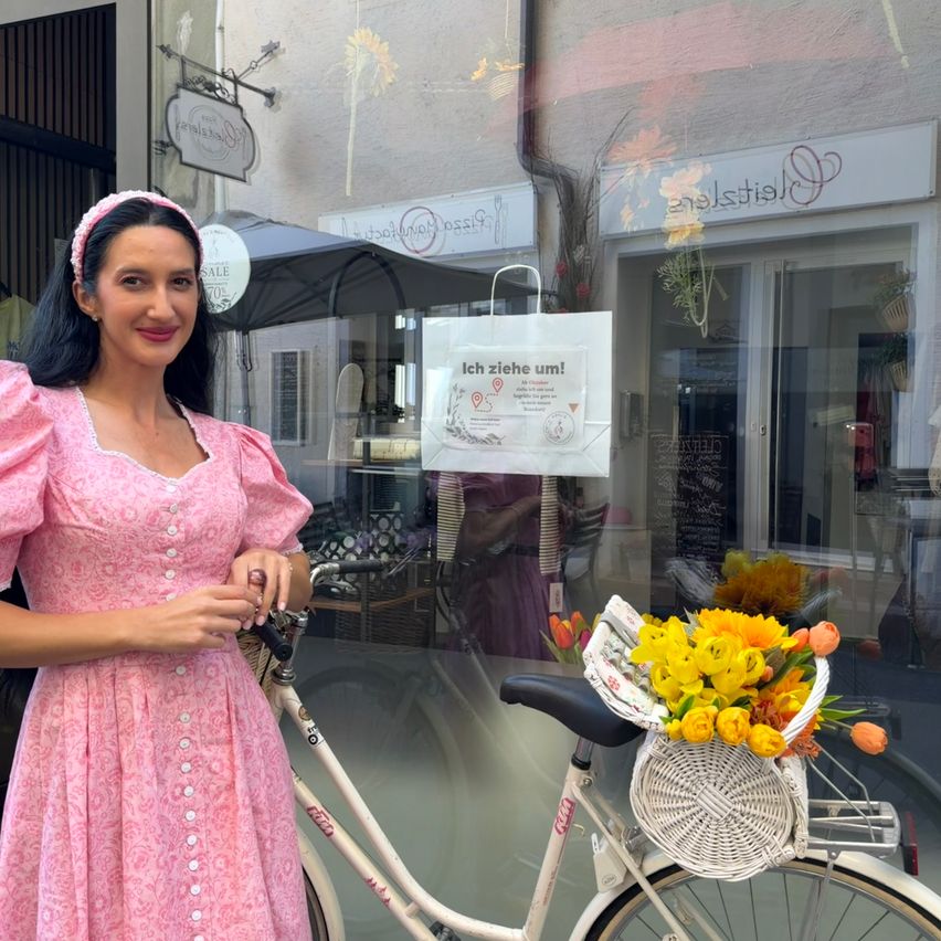 Eine Frau in einem rosa Kleid steht neben einem Fahrrad mit einem Korb gelber Blumen. Hinter ihr befindet sich ein Geschäft mit einem Schild, das 'Ich ziehe um!' besagt, und einer Reflexion einer Person.
