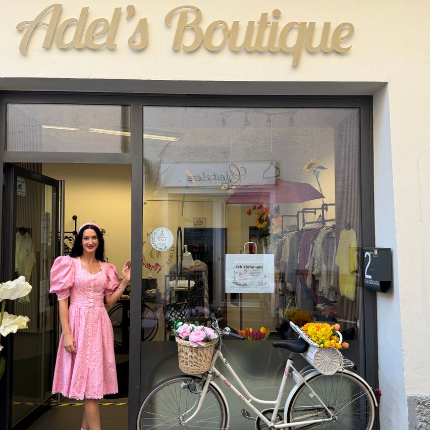 Eine Frau in einem rosa Kleid steht vor einem Boutique namens Adel's Boutique, mit einem Fahrrad mit Blumen im Vordergrund.