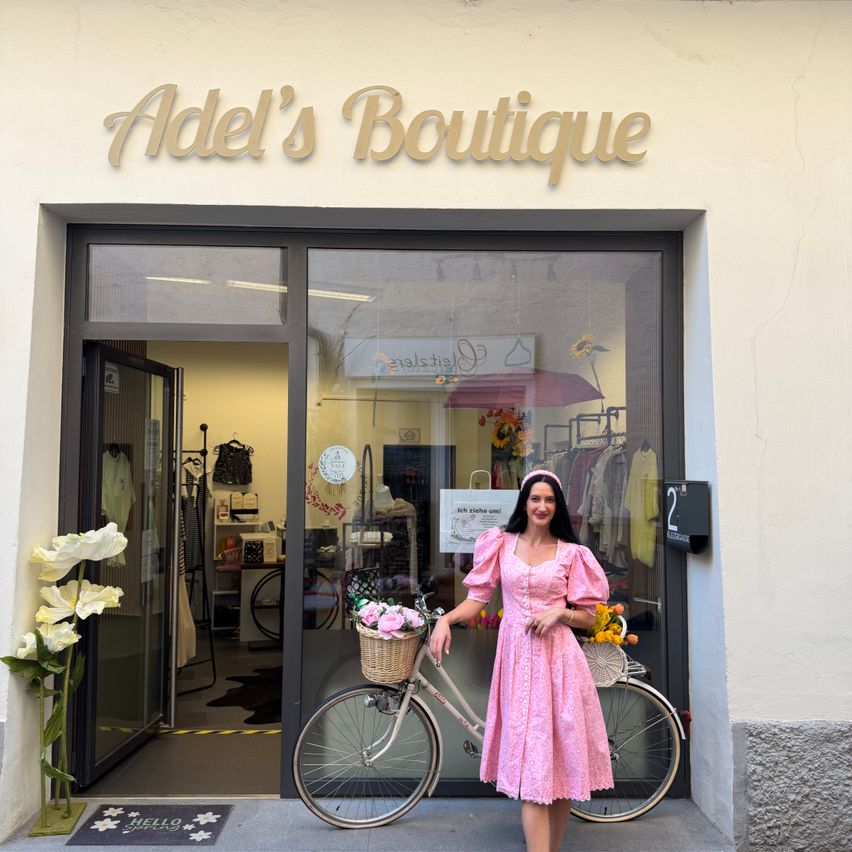 Eine Frau in einem rosa Kleid steht vor Adel's Boutique, hält ein Fahrrad mit einem Korb voller Blumen. Der Eingang des Ladens ist offen und zeigt eine Ausstellung von Kleidung im Inneren.