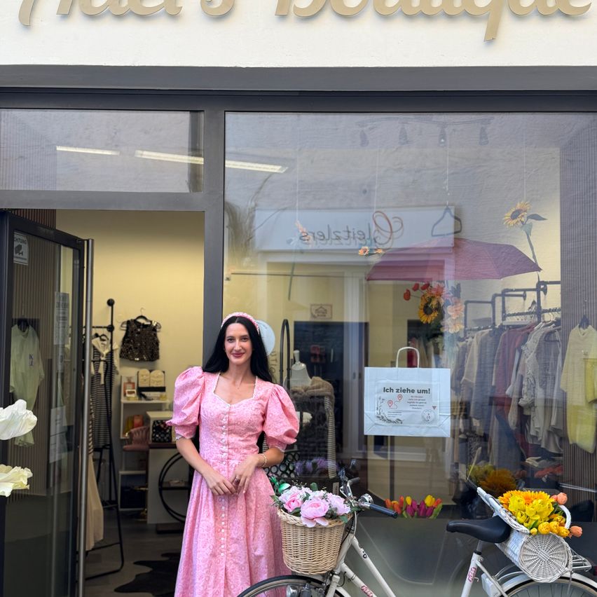 Eine Frau in einem rosa Kleid steht vor einem Boutique, hält ein Fahrrad mit einem Korb voller Blumen. Das Ladenschild lautet 'Mais Boutique'.