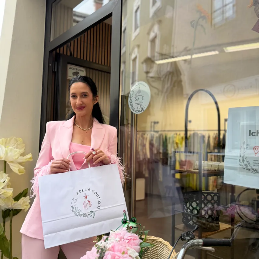 Eine Frau in einem rosa Anzug steht vor einem Boutique, hält einen Einkaufsbeutel mit Logo und lächelt in die Kamera.