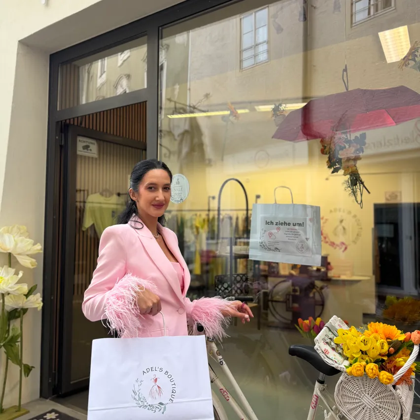 Eine Frau in einem rosa Anzug steht vor einem Geschäft und hält einen Einkaufsbeutel und ein Fahrrad. Im Schaufenster sind Blumen und ein Schild zu sehen.