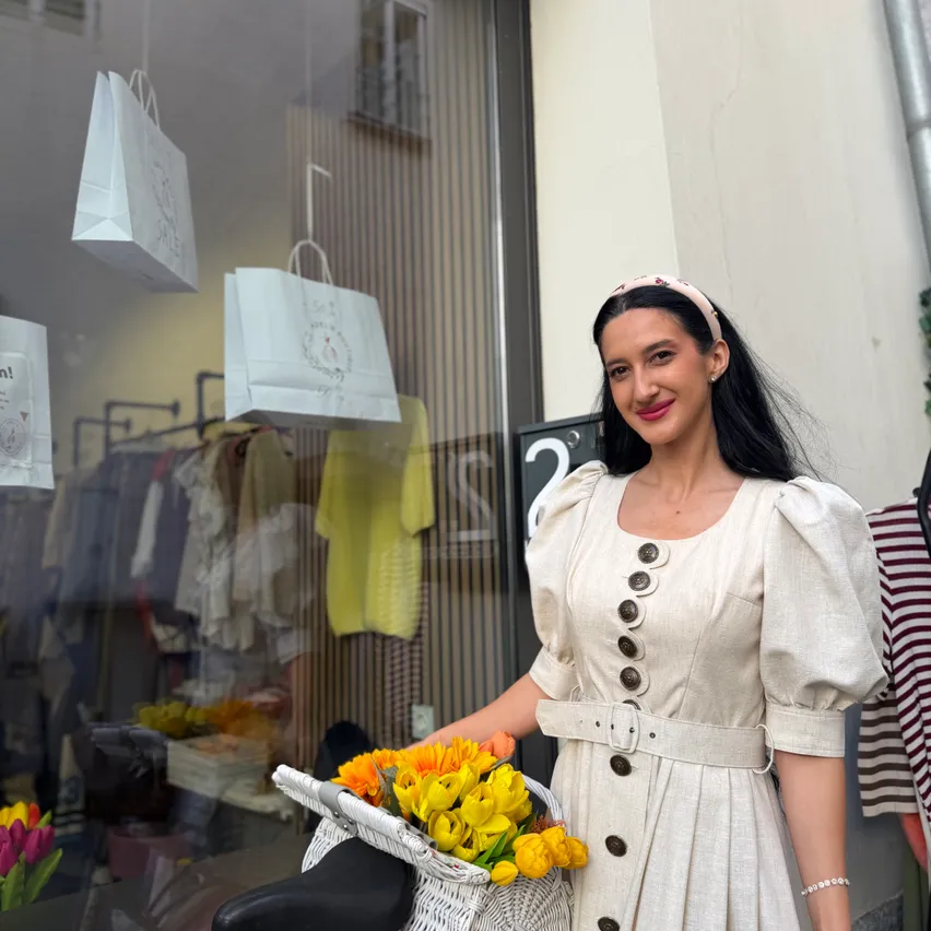 Eine Frau in einem weißen Kleid hält einen Korb mit gelben Blumen vor einem Boutique.