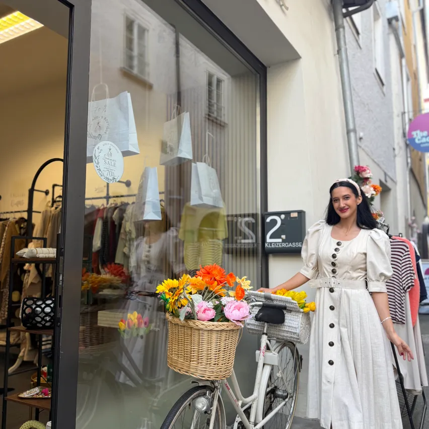 Eine Frau in einem weißen Kleid steht neben einem Fahrrad mit einem Blumenkorb vor einem Geschäft.