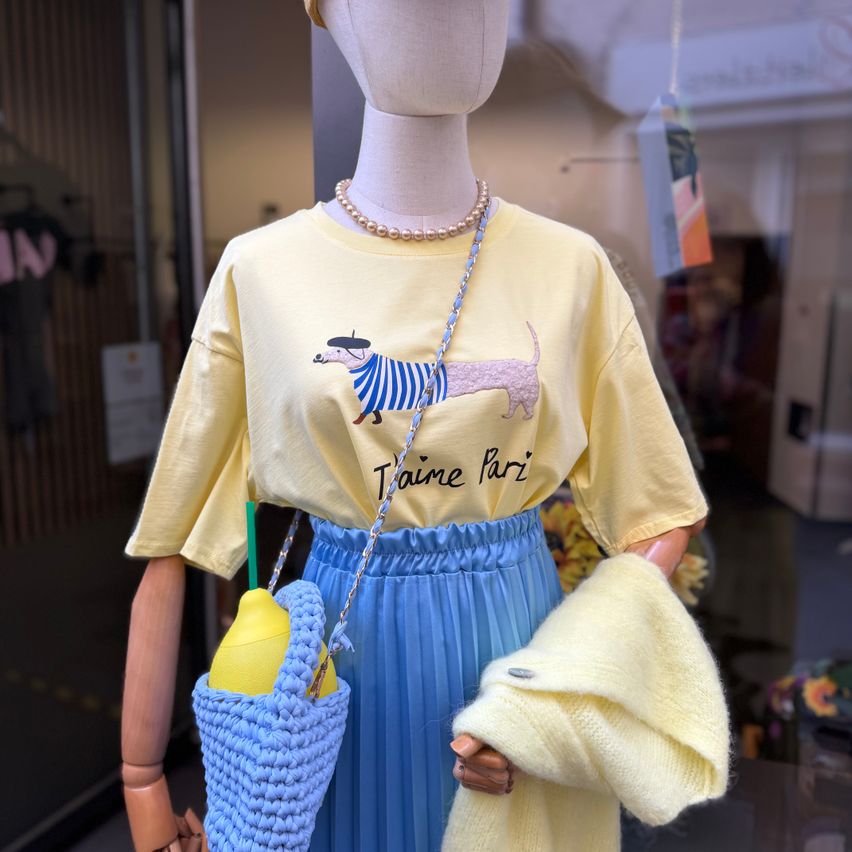 Eine Puppe im Schaufenster eines Ladens, die ein gelbes T-Shirt mit Dackeldesign, eine Perlenkette, einen blauen Rock und einen gelben gestrickten Cardigan mit einer blauen gehäkelten Tasche trägt.