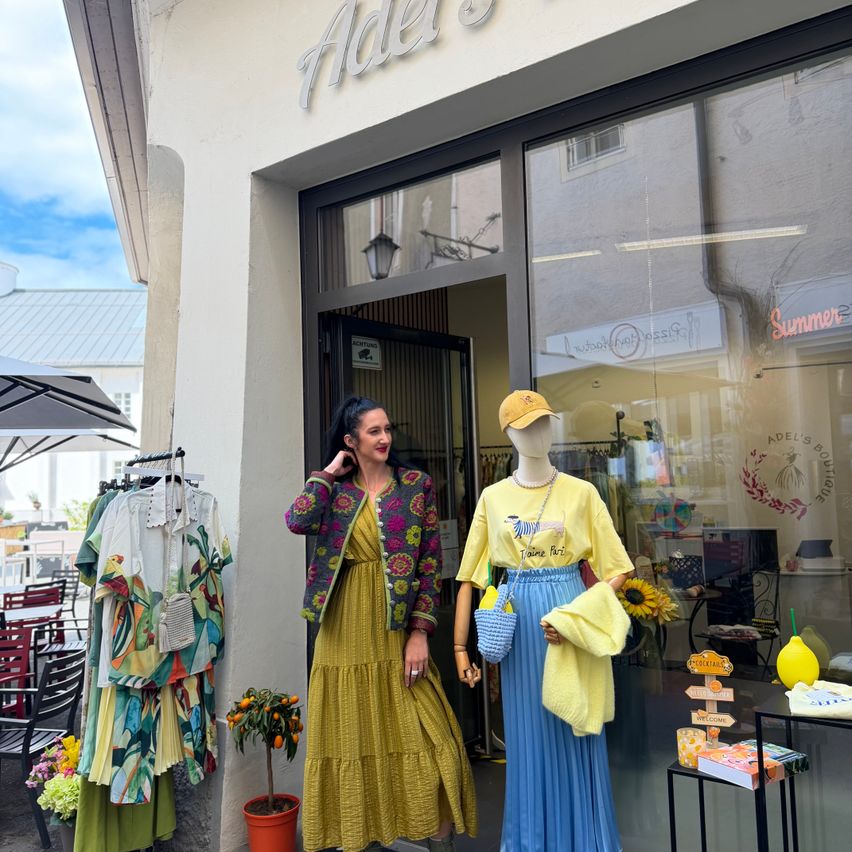 Eine Frau steht vor dem Adel's Boutique, zeigt ein leuchtend gelbes Kleid und eine blumige Strickjacke. Ein Mannequin in einem passenden gelben Oberteil und einem blauen Rock steht neben ihr, mit einem Regenschirm und Blumen im Vordergrund.