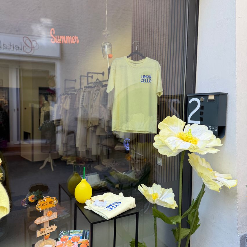 Ein Schaufenster zeigt ein gelbes Limon Cello T-Shirt, mit künstlichen Blumen und einer Zitrone mit Strohhalm. Im Inneren hängen Kleider, und es gibt ein 'Cocktails' Schild.