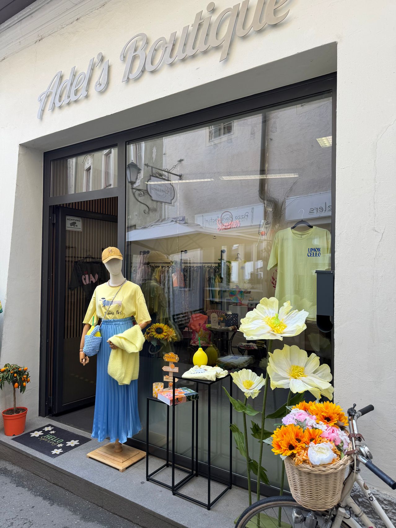 Ein Boutique-Schaufenster mit einer Puppe, die ein gelbes Hemd und eine blaue Rock trägt. Das Fenster zeigt eine Blumenarrangement und ein gelbes T-Shirt. Der Name des Ladens lautet Adel's Bar.
