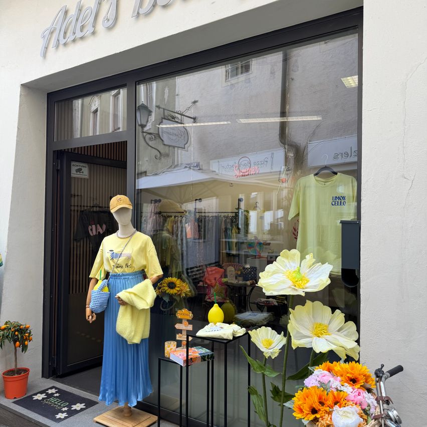 Ein Boutique-Schaufenster mit einer Puppe, die ein gelbes Hemd und eine blaue Rock trägt. Das Fenster zeigt eine Blumenarrangement und ein gelbes T-Shirt. Der Name des Ladens lautet Adel's Bar.