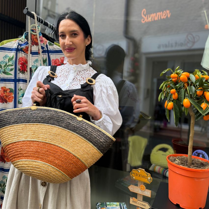 Eine Frau in traditioneller Kleidung steht vor einem Schaufenster und hält eine bunte gewebte Tasche. Dahinter ist ein Topf mit Orangenbaum und ein Schild mit der Aufschrift 'Cocktails' zu sehen.