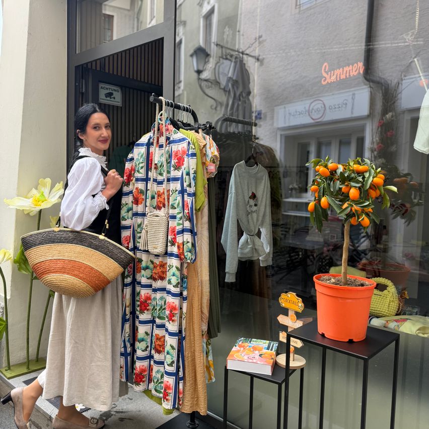 Eine Frau steht vor einem Bekleidungsgeschäft, hält einen Korb und eine Tasche und Kleidungsstücke hängen an einem Bügel im Inneren. Ein Topf mit Pflanzen und Bücher sind auf einem Tisch im Schaufenster ausgestellt.