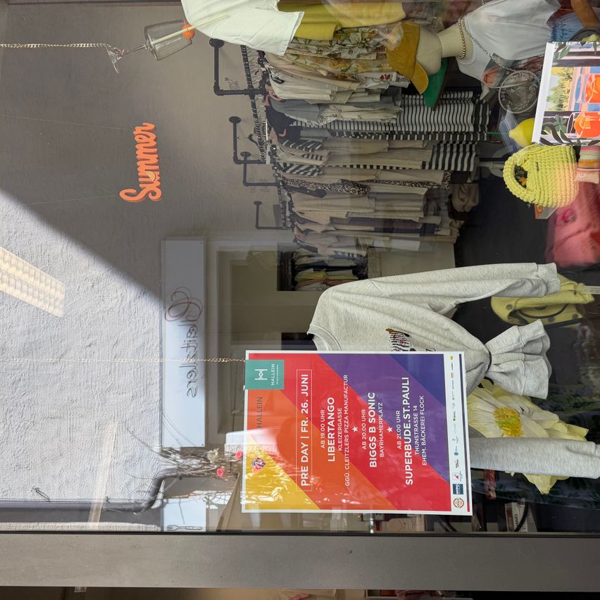 Ein Geschäft zeigt verschiedene Kleidungsstücke. Ein Schild bewirbt eine Veranstaltung am 26. Juni. Das Schild enthält Bandnamen, Zeiten und einen Ort. Das Wort 'Sommer' ist an der Wand sichtbar.