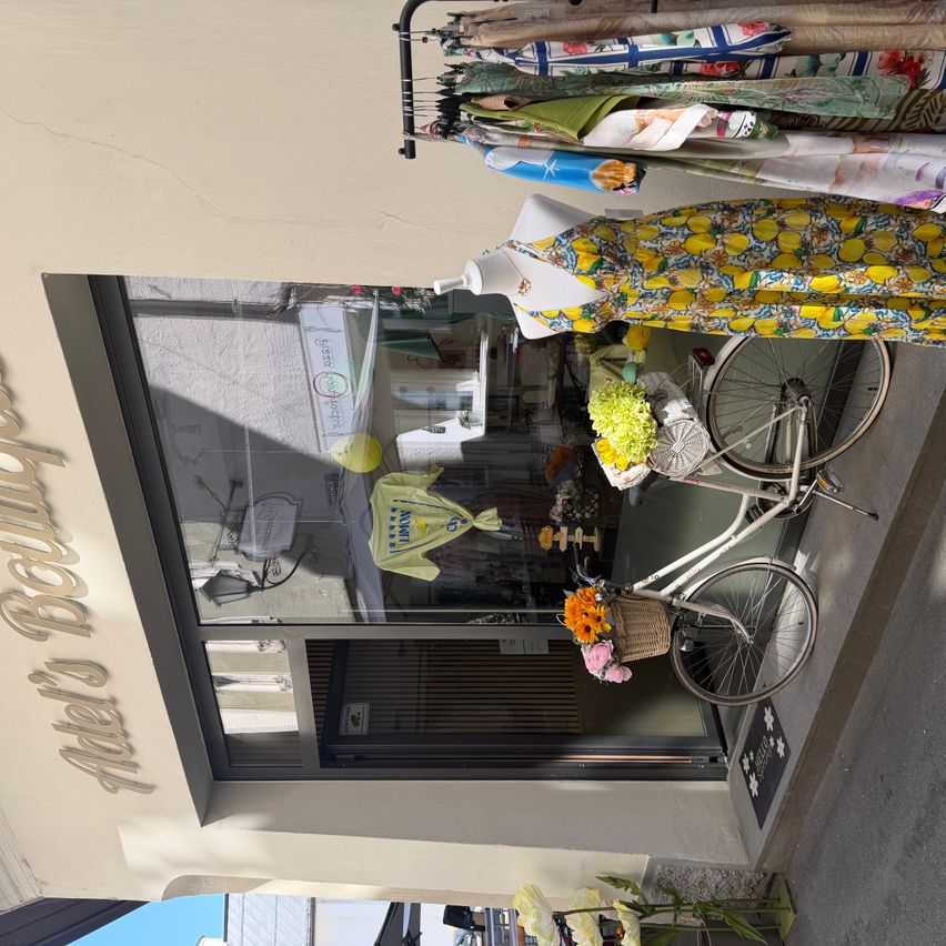 Ein Blick von oben auf eine Boutique. Ein weißes Fahrrad mit Blumen im Korb ist draußen geparkt. Das Geschäft hat ein großes Glasfenster, das verschiedene Kleidungsstücke zeigt. Der Name des Geschäfts, 'Boutique Bella', ist an der Wand geschrieben.