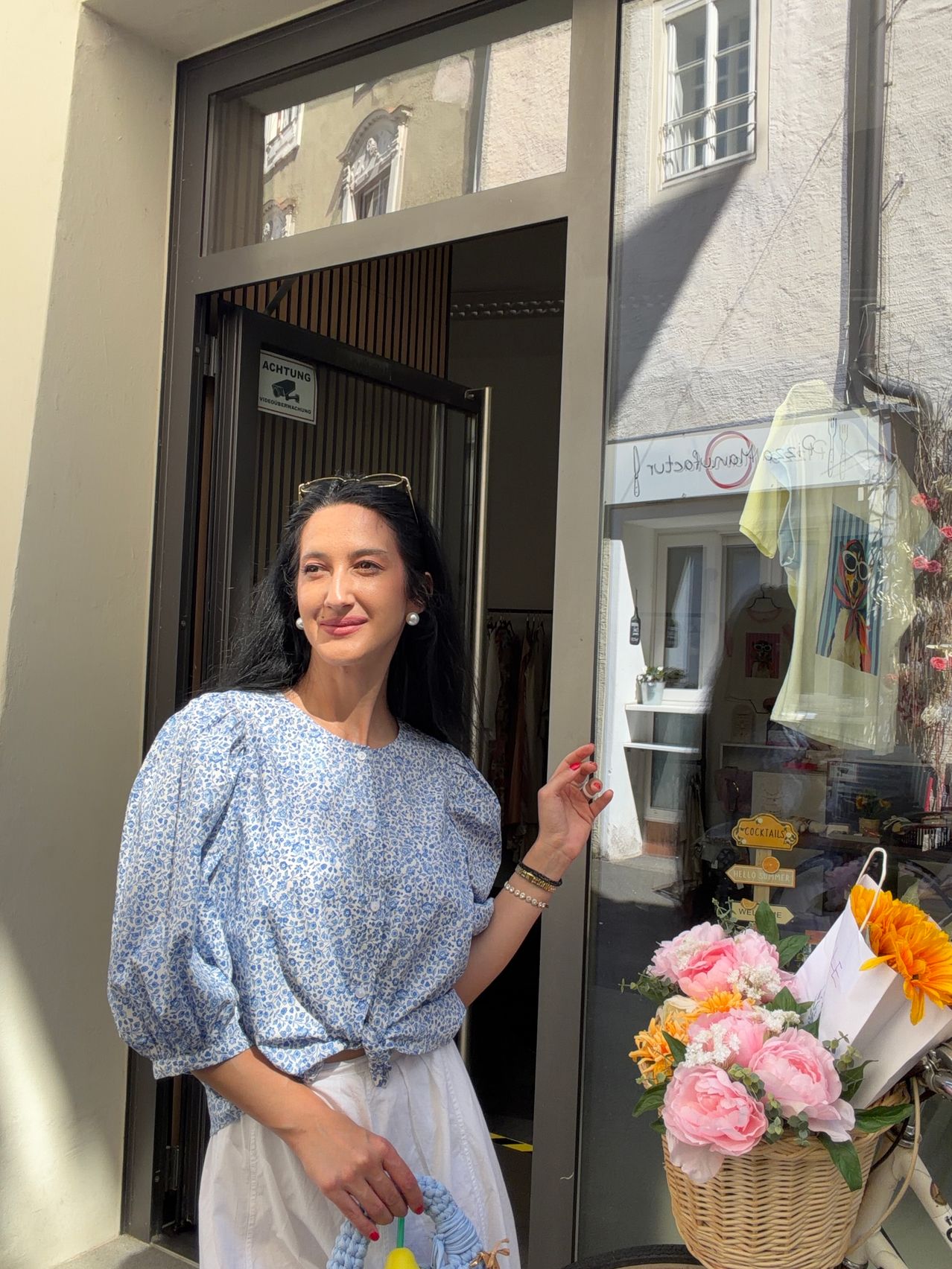 Eine Frau steht vor einem Boutique, lächelt, mit einem Blumenstrauß und einem Schild, das 'ACHTUNG' über der Tür steht.