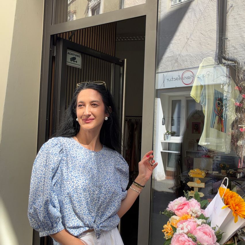 Eine Frau steht vor einem Boutique, lächelt, mit einem Blumenstrauß und einem Schild, das 'ACHTUNG' über der Tür steht.