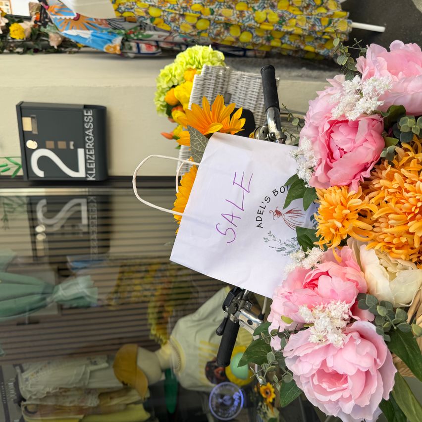 Ein Fahrrad mit einem Korb voller Blumen ist vor einem Laden geparkt. Ein Papierbeutel mit dem Wort SALE ist am Korb befestigt. Der Name des Ladens ist Kleinergasse.