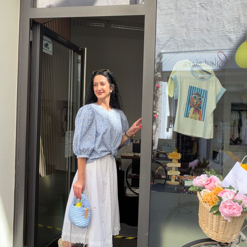 Eine Frau steht vor einem Geschäft, lächelt. Sie trägt ein blaues Oberteil und eine weiße Rock, hält einen Korb. Das Schaufenster zeigt ein Hunde-T-Shirt, Blumen und ein Fahrrad.