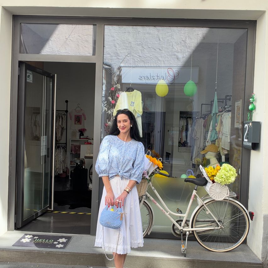 Eine Frau steht vor einem Boutique-Laden mit einem Fahrrad davor. Sie trägt ein blaues Oberteil und einen weißen Rock. Der Laden zeigt Kleidung und bunte Ballons im Inneren.
