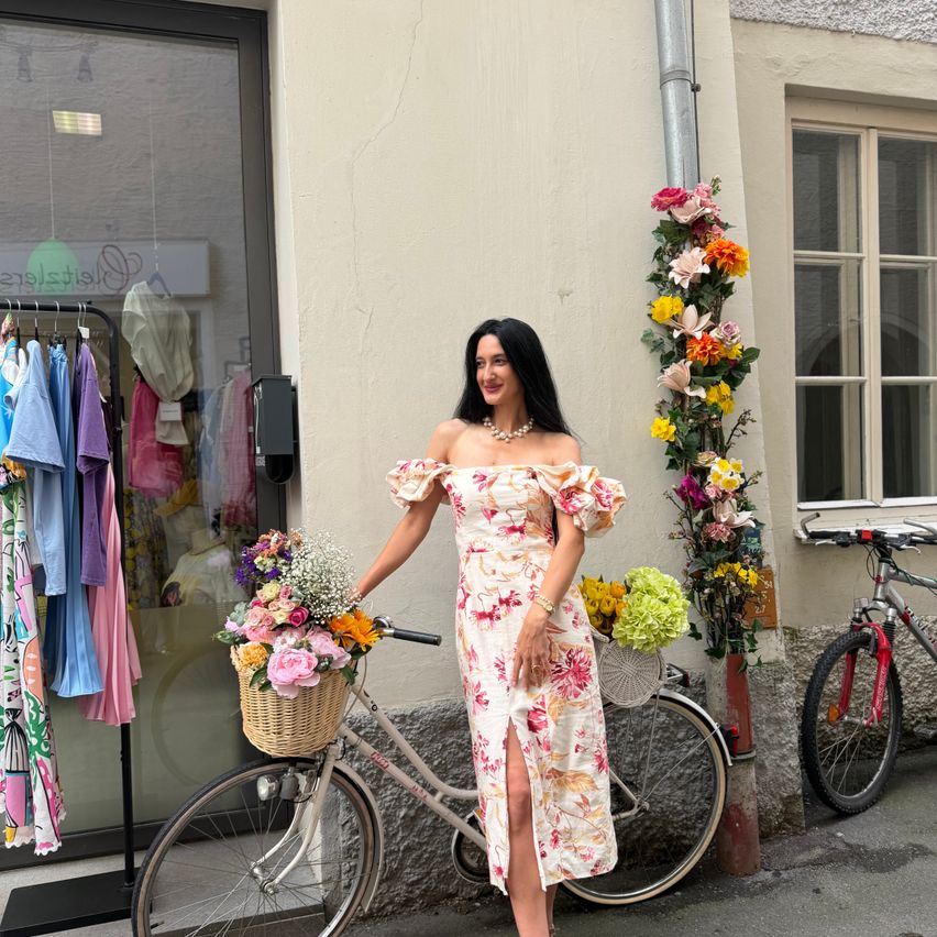 Eine Frau steht neben einem Fahrrad mit einem Korb voller Blumen vor einem Bekleidungsgeschäft.