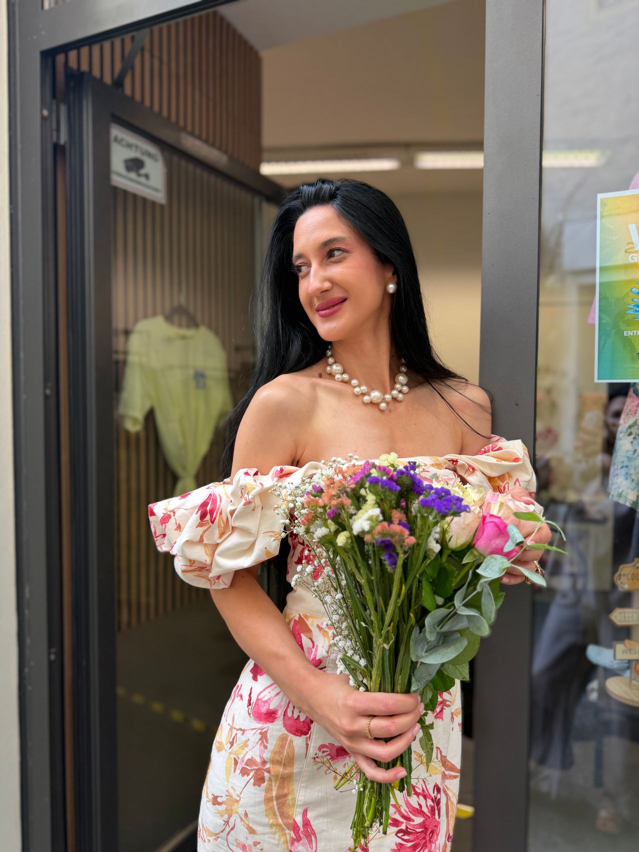 Eine Frau mit schwarzen Haaren, lächelnd, steht in einem Türrahmen und hält einen Blumenstrauß. Sie trägt eine Perlenkette und ein Blumenkleid. Hinter ihr sind ein Schild und Kleidung sichtbar.