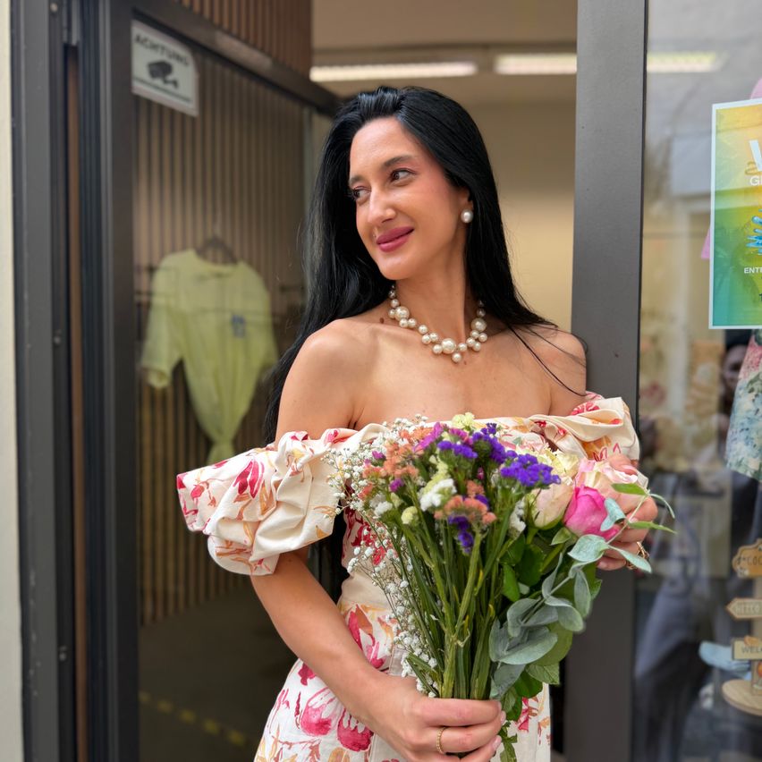 Eine Frau mit schwarzen Haaren, lächelnd, steht in einem Türrahmen und hält einen Blumenstrauß. Sie trägt eine Perlenkette und ein Blumenkleid. Hinter ihr sind ein Schild und Kleidung sichtbar.
