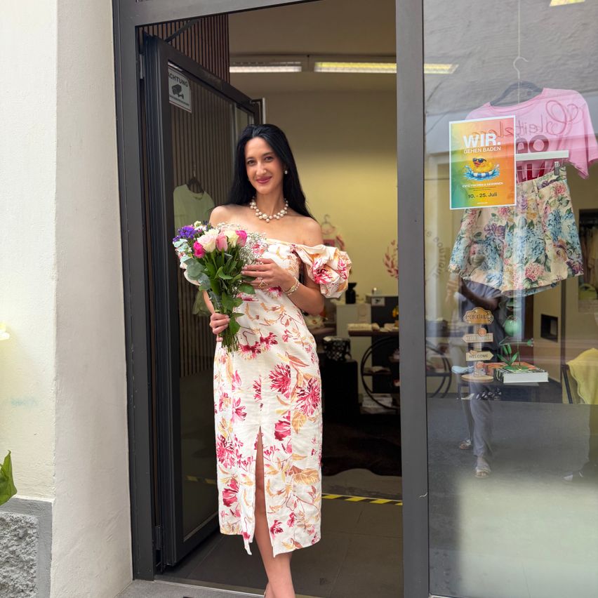 Eine Frau mit Blumen steht vor einem Ladeneingang und lächelt für ein Foto. Hinter ihr hängen Kleidungsstücke an Kleiderbügeln. Der Laden hat eine Glastür und ein Schild mit Text und einem gelben Plakat.