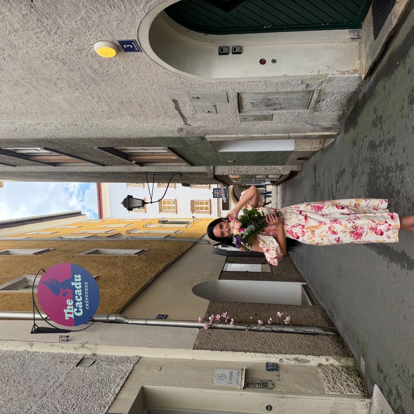 Eine Frau in einem Blumenkleid hält einen Blumenstrauß und posiert für ein Foto in einer engen Straße zwischen Gebäuden.