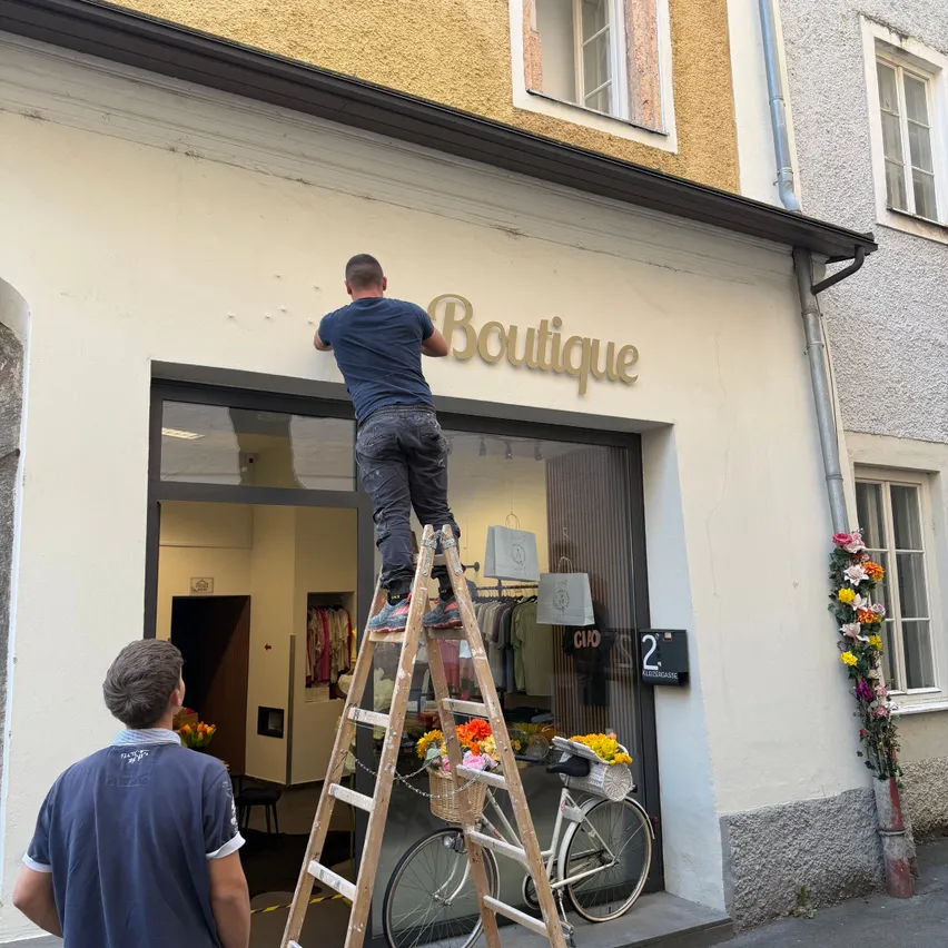 Ein Mann steht auf einer Leiter und malt das Schild einer Boutique an einem Gebäude.