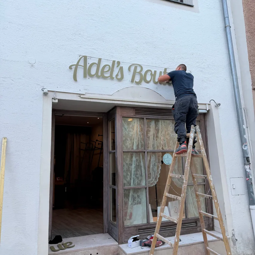 Ein Mann steht auf einer Leiter vor einem Geschäft und repariert ein Schild, das Adel's Boutique liest.