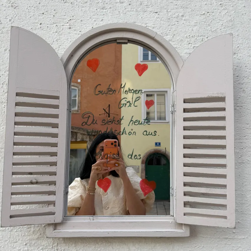 Eine Frau macht ein Selfie vor einem Fenster mit einem herzförmigen Design und einer deutschen Botschaft darauf.