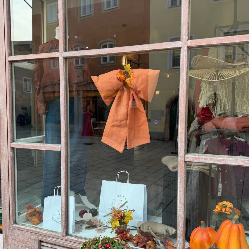 Ein Schaufenster mit einem orangefarbenen Bogen und Herbstdekorationen. Im Inneren befinden sich weiße Einkaufstüten, eine Mütze und Kleidung. Draußen ist die Reflexion eines Gebäudes und einer Person zu sehen.