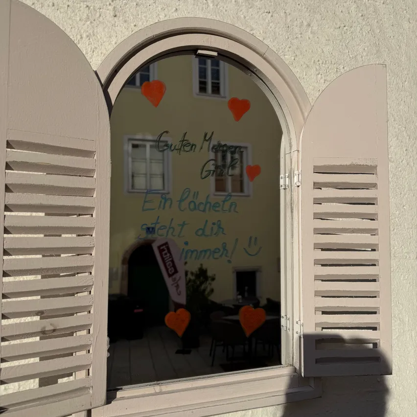 Ein Fenster mit weißen Fensterläden und einer auf dem Glas geschriebenen Nachricht auf Deutsch.