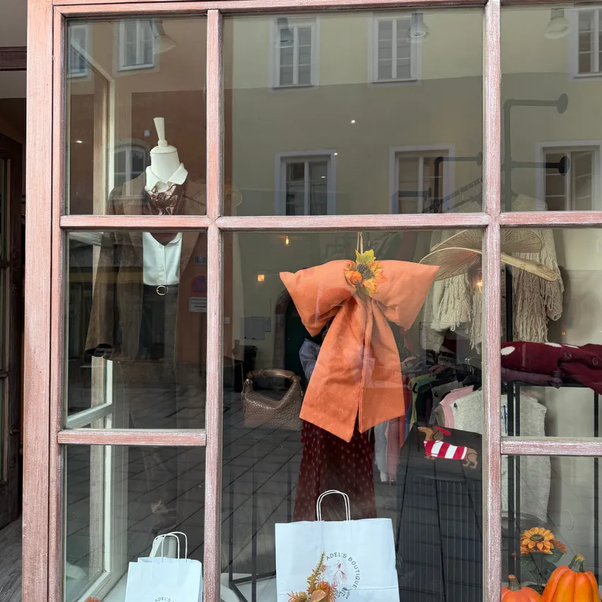 Ein Schaufenster zeigt eine Puppe, die eine braune Jacke und ein weißes Hemd trägt. Im Fenster befinden sich ein großer orangefarbener Bogen, ein weißer Papierbeutel mit Logo und verschiedene Gegenstände wie Kürbisse, Blätter und eine kleine Pflanze.