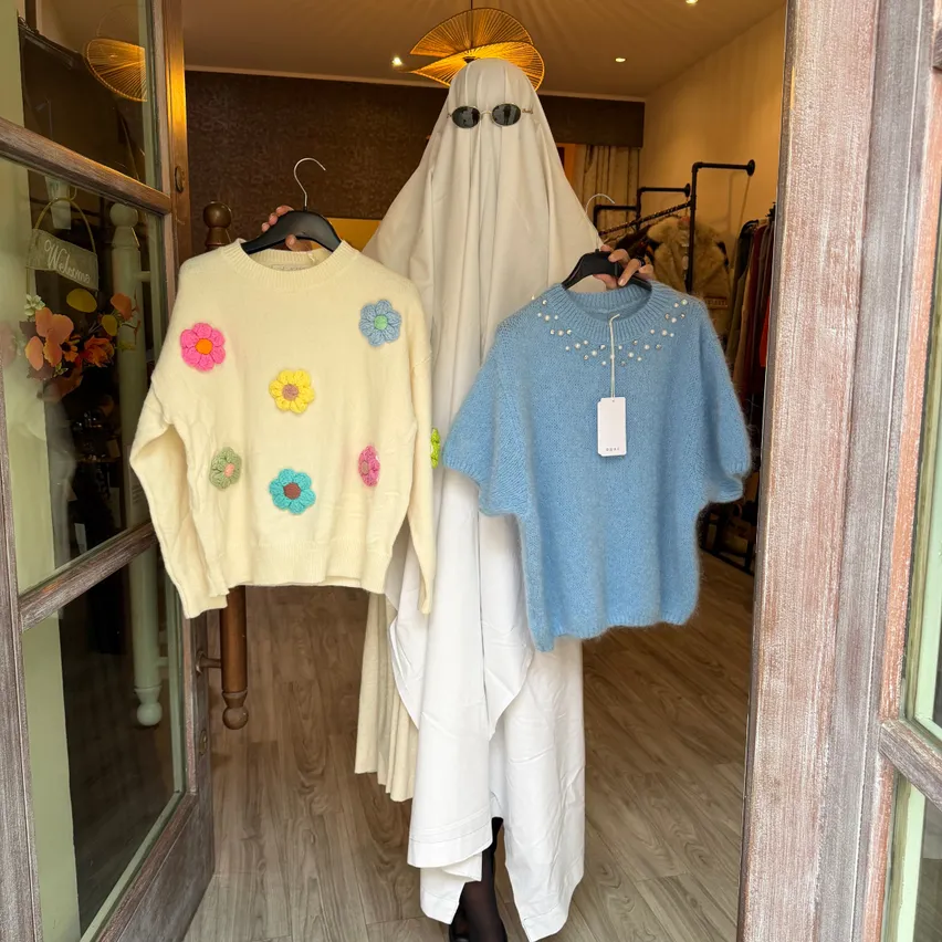 Eine Person in einem Geisterkostüm, die zwei Pullover in einem Bekleidungsgeschäft hält.