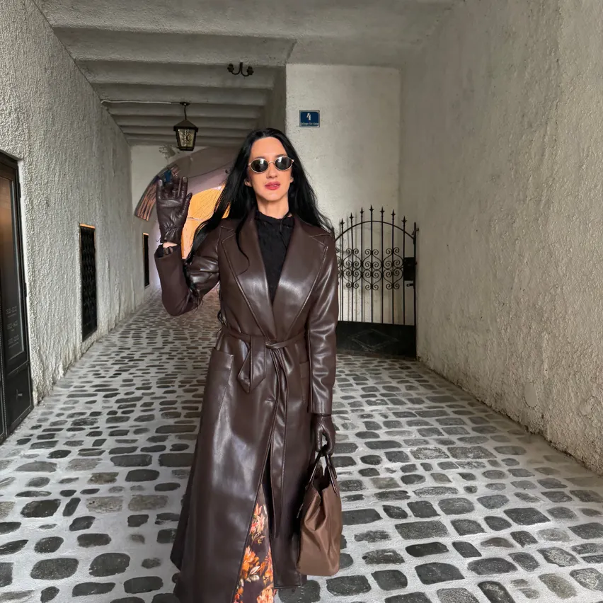 Eine Frau in einem braunen Ledermantel steht in einer Kopfsteinpflastergasse und posiert für ein Foto.