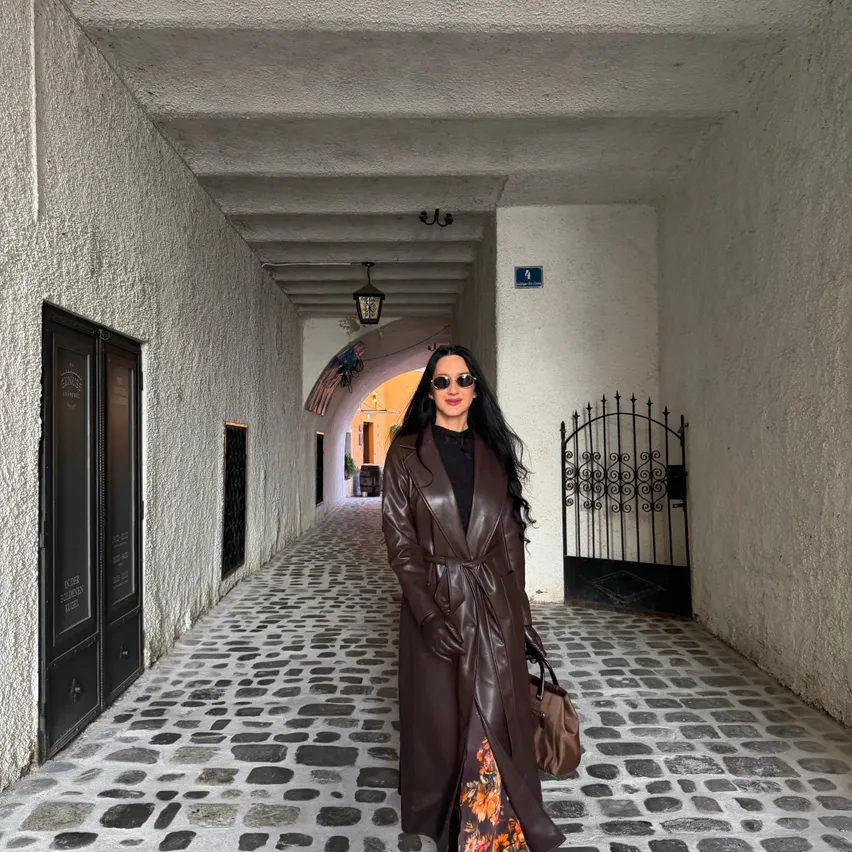 Eine Frau in einem braunen Ledermantel und einem Blumenrock steht in einer Kopfsteinpflastergasse mit weißen Wänden und einem schwarzen Tor.